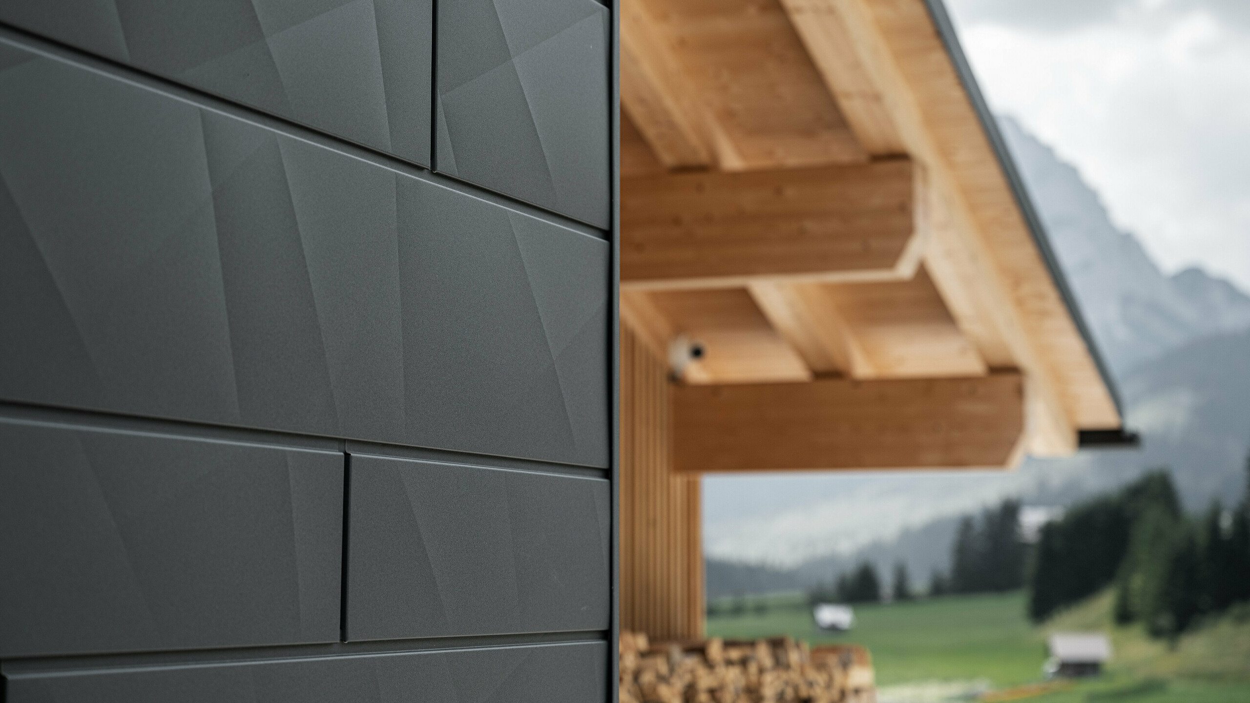 Nahaufnahme der Siding.X Fassadenpaneele in P.10 Schwarz von PREFA an der Casa Hoffe in Sappada, Italien. Das Fassadensystem aus Aluminium ist gekonnt in einem modernen, abstrakten Design angebracht, das einen ansprechenden Kontrast zu den sichtbaren Holzelementen des Dachstuhls bildet. Die innovative Fassadenlösung fügt sich nahtlos in das alpine Ambiente ein und steht für die Verbindung von Funktionalität, Beständigkeit und ästhetischer Baukultur.