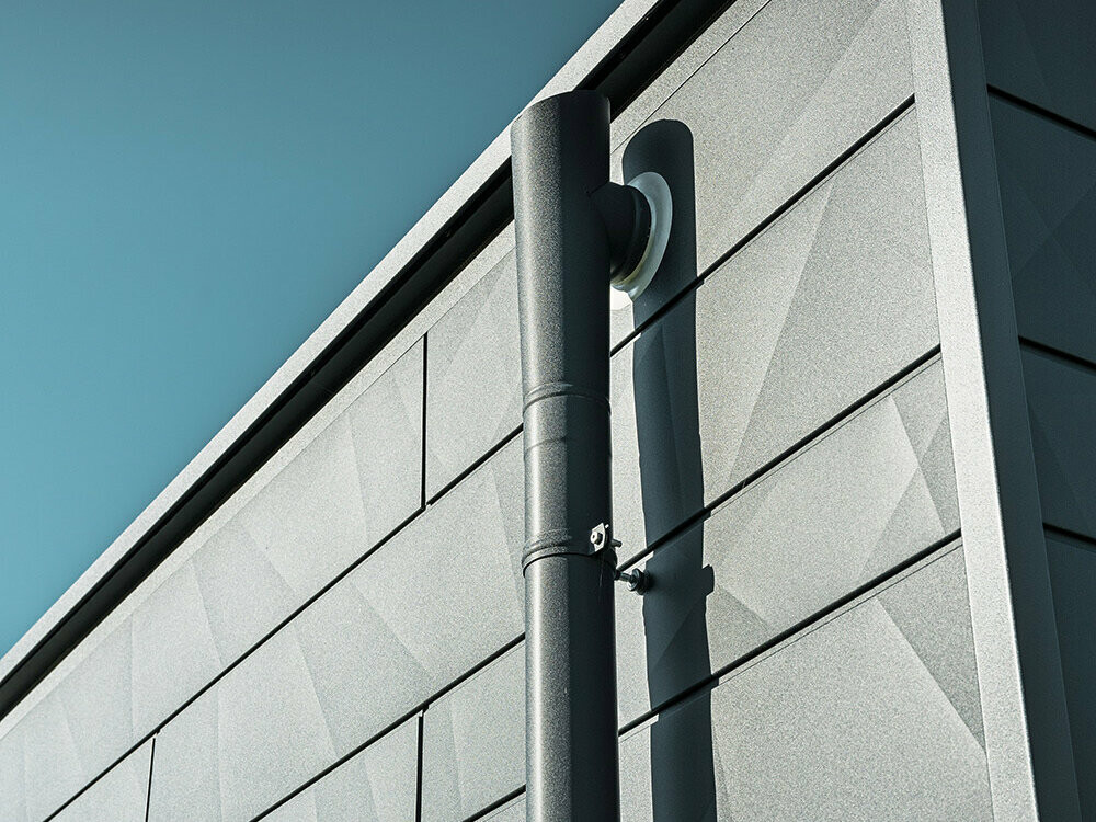 PREFA Ablaufrohr mit PREFA Speiereinmüngung in P.10 Anthrazit PREFA Ablaufrohr mit PREFA Speiereinmüngung in P.10 Anthrazit an einer PREFA Siding.X Fassade, ebenfalls in P.10 Anthrazit