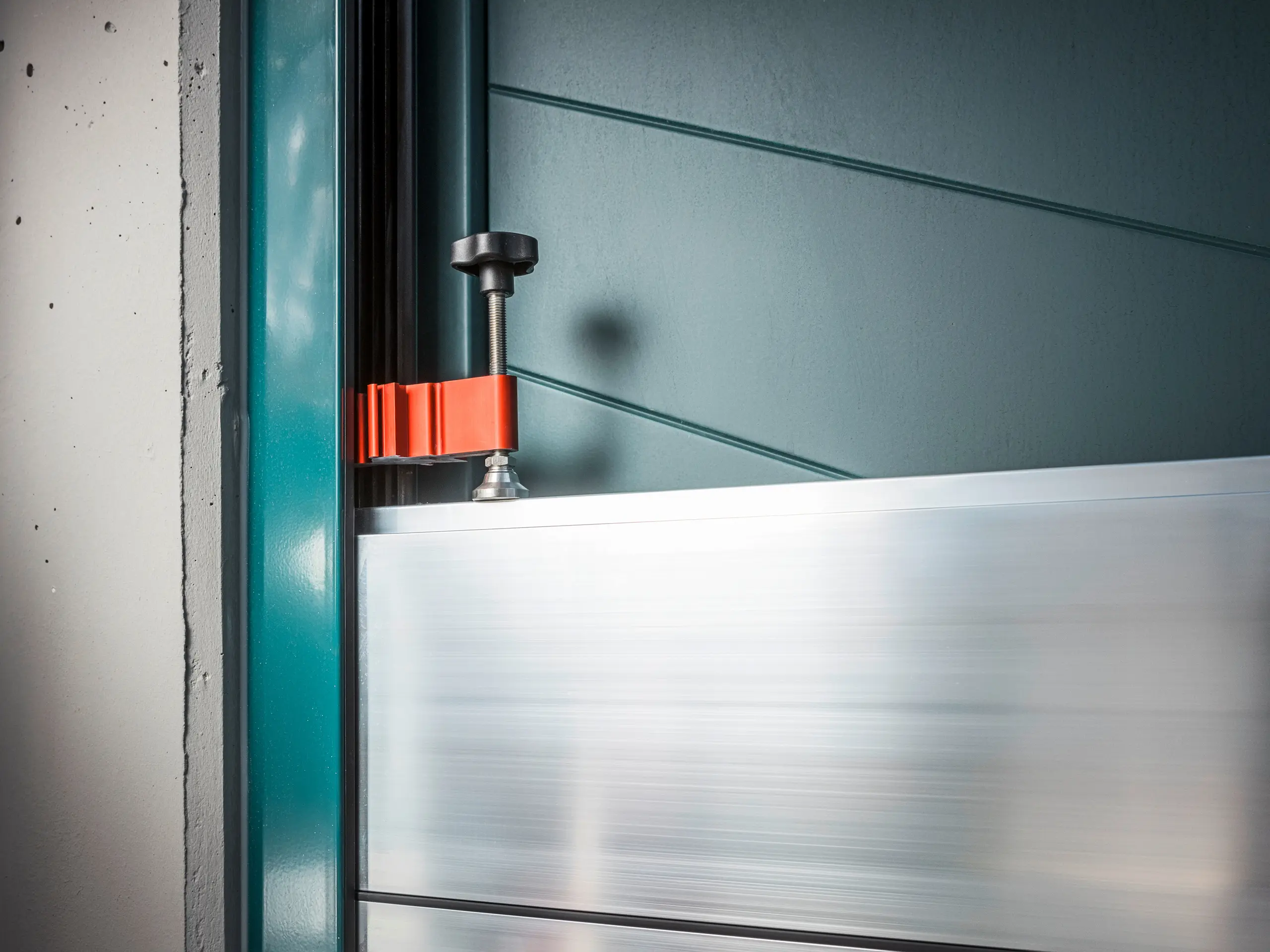 Detailaufnahme des PREFA Hochwasserschutzsystems an einem Torrahmen, bestehend aus Aluminium-Dammbalken, die mit einer robusten, roten Klemme (dem Spannstück) befestigt sind, um sicheren Schutz vor Hochwasser zu gewährleisten.