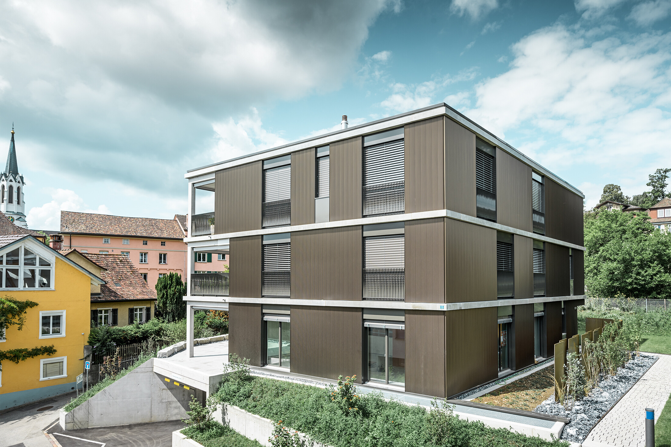 Die markante Aluminiumfassade dieses Mehrfamilienhauses in Schaffhausen ist mit dem eleganten PREFA Zackenprofil in der Farbe Colinal ausgestattet. Durch die vertikale Anordnung der stranggepressten Aluminiumprofile wirkt das Gebäude optisch kompakter und die Fassadenstruktur erhält eine harmonische Balance. Das edle Design fügt sich geschmackvoll in die Umgebung ein und verleiht dem Gebäude eine moderne Eleganz, die sowohl optisch ansprechend als auch funktional ist. Die Perspektive zeigt das Mehrfamilienhaus von der Vorderseite mit der Garageneinfahrt. Im Hintergrund sind Bäume, weitere Gebäude und ein Kirchturm zu erkennen.