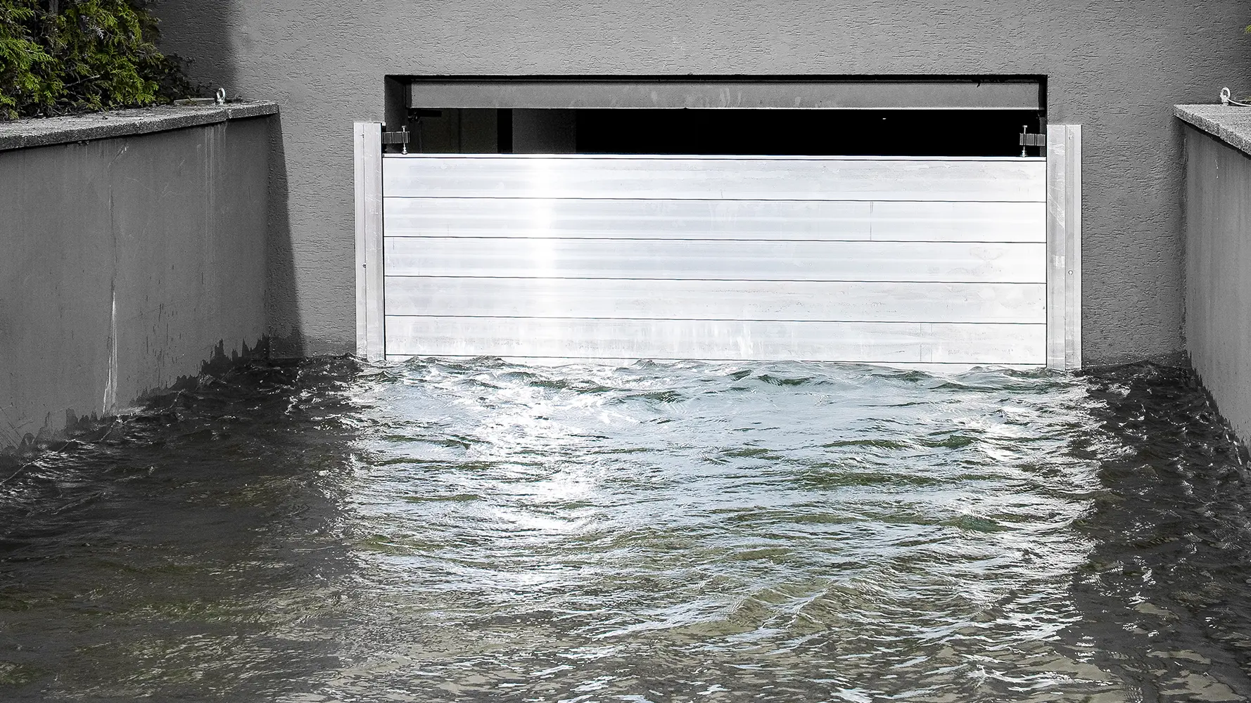 PREFA Hochwasserschutz in Aktion: Die Aluminium-Barriere schützt effektiv gegen Hochwasser. Die glänzende Metallwand trotzt dem aufgewühlten Wasser und schützt die dahinterliegende Garage. Diese HWS-Lösung verbindet Funktionalität mit einem modernen, ästhetischen Design und demonstriert nachhaltigen Objekt- und Landschaftschutz mit der Stärke von PREFA.