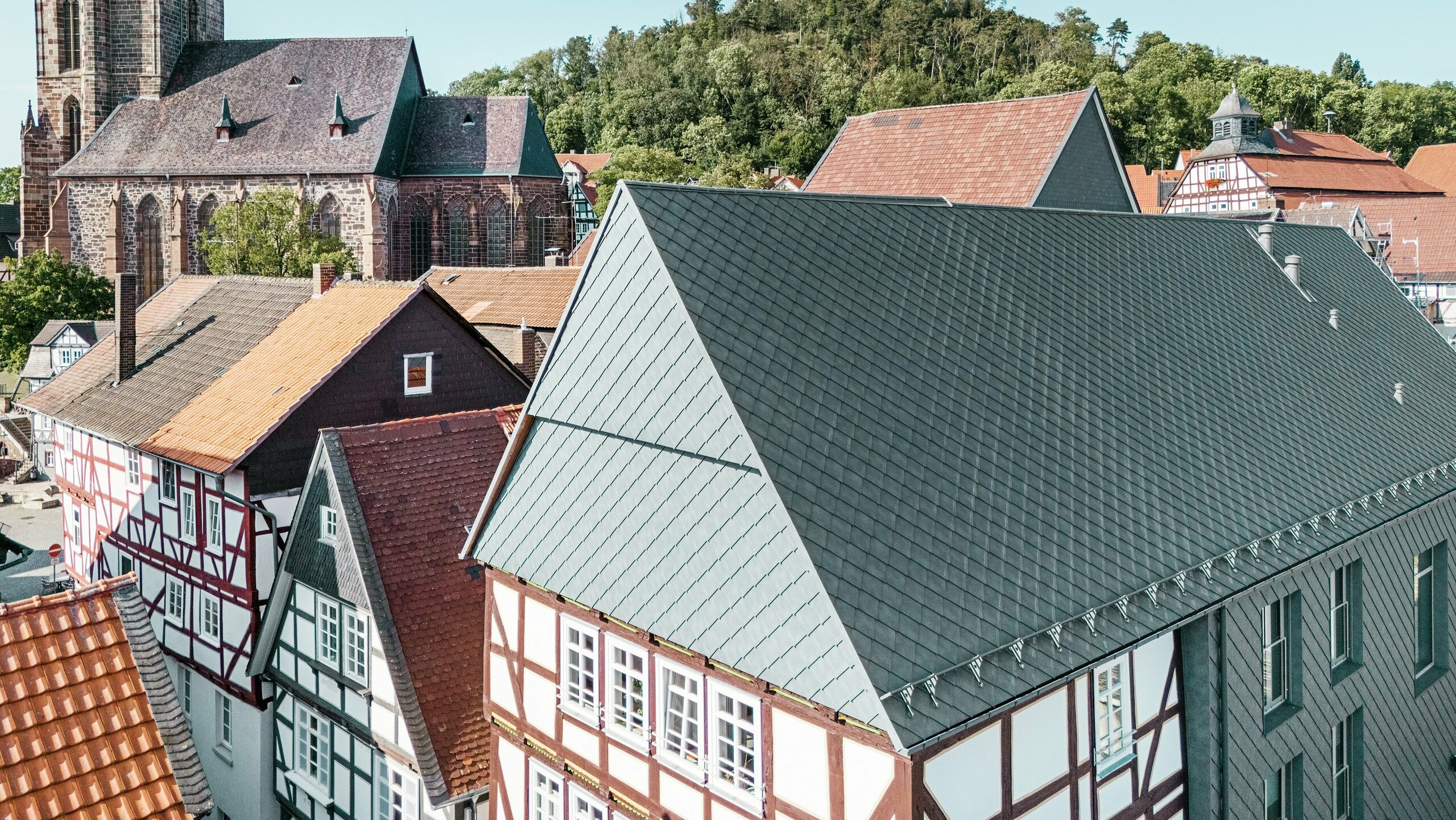 The Volkshaus in Homberg, Germany Das Dach des sanierten Volkshauses in Homberg, DE, mit PREFA Aluminiumrauten 29 × 29 in P.10 Steingrau, eingebettet in die historische Altstadt mit Fachwerkhäusern, Kirche und Burg. Die langlebige und robuste Aluminiumverkleidung verbindet modernes Design mit dem Charme der historischen Umgebung. In der Bildmitte ist der Giebel des Gebäudes zu sehen, im Hintergrund die Kirche und der Burgberg.