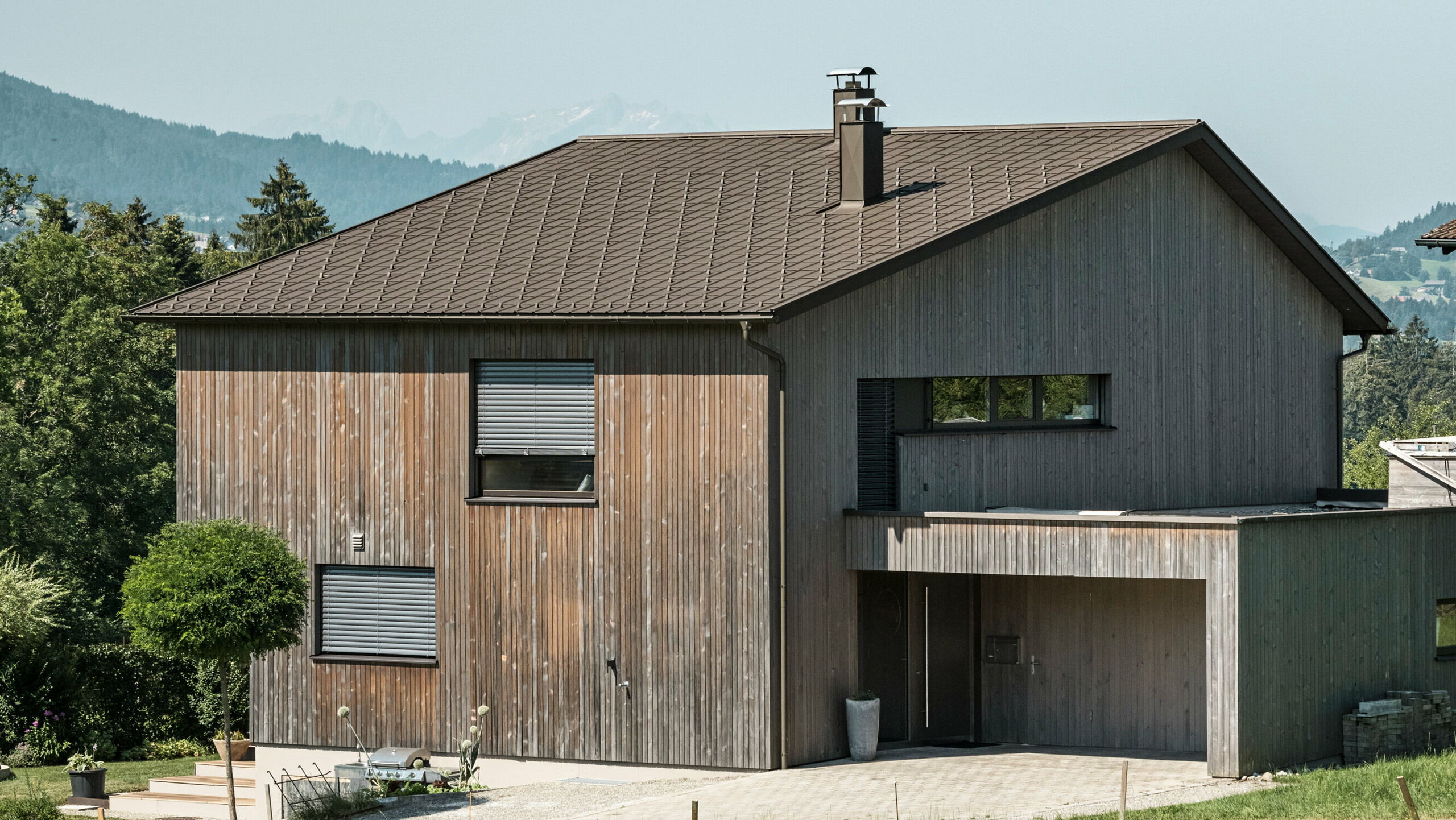 Wooden detached house with PREFA roof tiles in P.10 brown Seitenansicht eines zeitgenössischen Einfamilienhauses in Krumbach mit einer Kombination aus einer natürlichen Holzfassade und einem langlebigen PREFA Aluminiumdach in P.10 Braun. Die harmonische Farbgebung zeigt die elegante Integration von modernem Design und traditionellen Materialien, wobei die PREFA Dachplatten für eine ästhetische und funktionale Dachgestaltung sorgen.