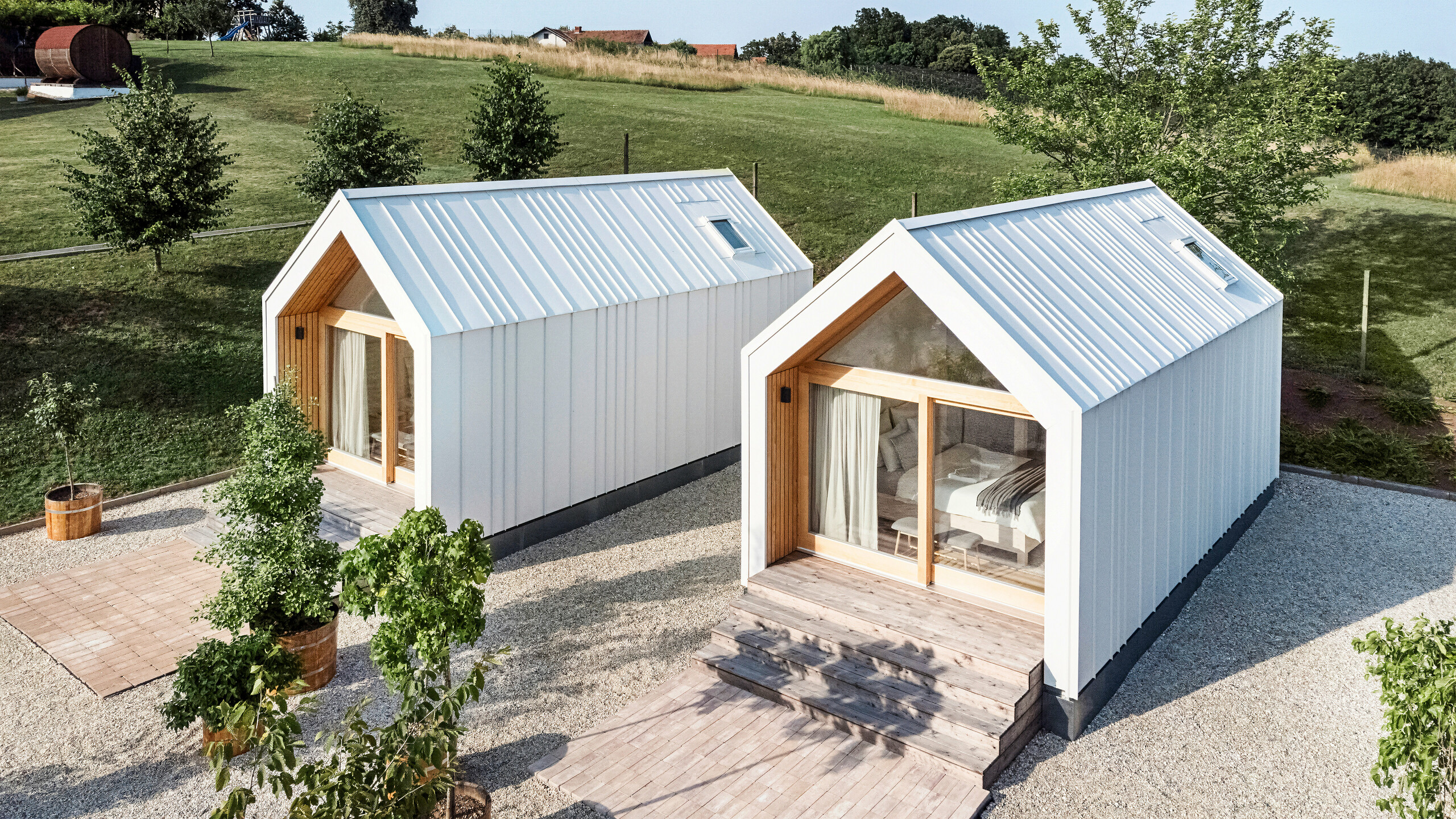 Tiny houses with PREFALZ roof and façade in P.10 pure white Blick auf die Vorderseite der beiden Tiny Häuser der Ferienanlage 'Pri Momi' in Kančevci, Slowenien. Die Mikrohäuser wurden in einer Holzrahmenbauweise erreichtet und mit einer robusten PREFALZ Dach- und Fassadenverkleidung in P.10 Reinweiß vollendet. Die modernen, minimalistischen Bauwerke kombinieren eine langlebige und zugleich ästhetische Aluminiumhülle mit großen Glasfronten und naturbelassenem Holz, eingebettet in eine grüne Landschaft. Die großzügigen Holzstufen führen zu den Eingängen, die eine gemütliche und einladende Atmosphäre schaffen.