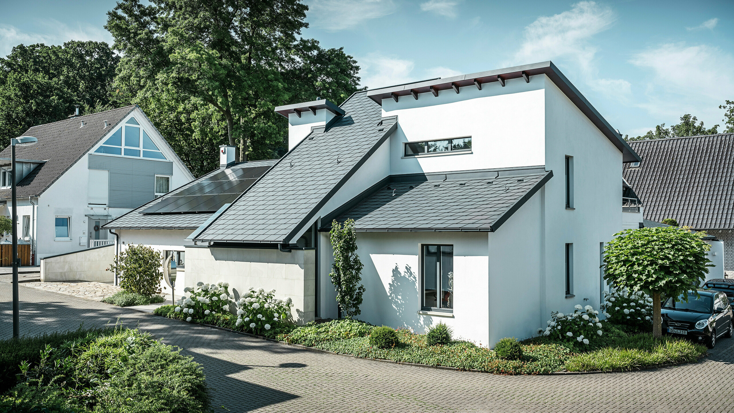 PREFA complete system: DS.19 roof shingle, Roof drainage and und Solar panel substructure for a single family house Modernes Einfamilienhaus mit weißer Putzfassade und dunklem PREFA Schrägdach aus Aluminium. Auf dem Dach sind integrierte Photovoltaikmodule und eine Aluminium-Dachentwässerung in Anthrazit zu sehen. Das Gebäude liegt in einer gepflegten Wohnsiedlung mit bepflanzten Vorgärten, gepflasterter Zufahrt und umliegenden Einfamilienhäusern.