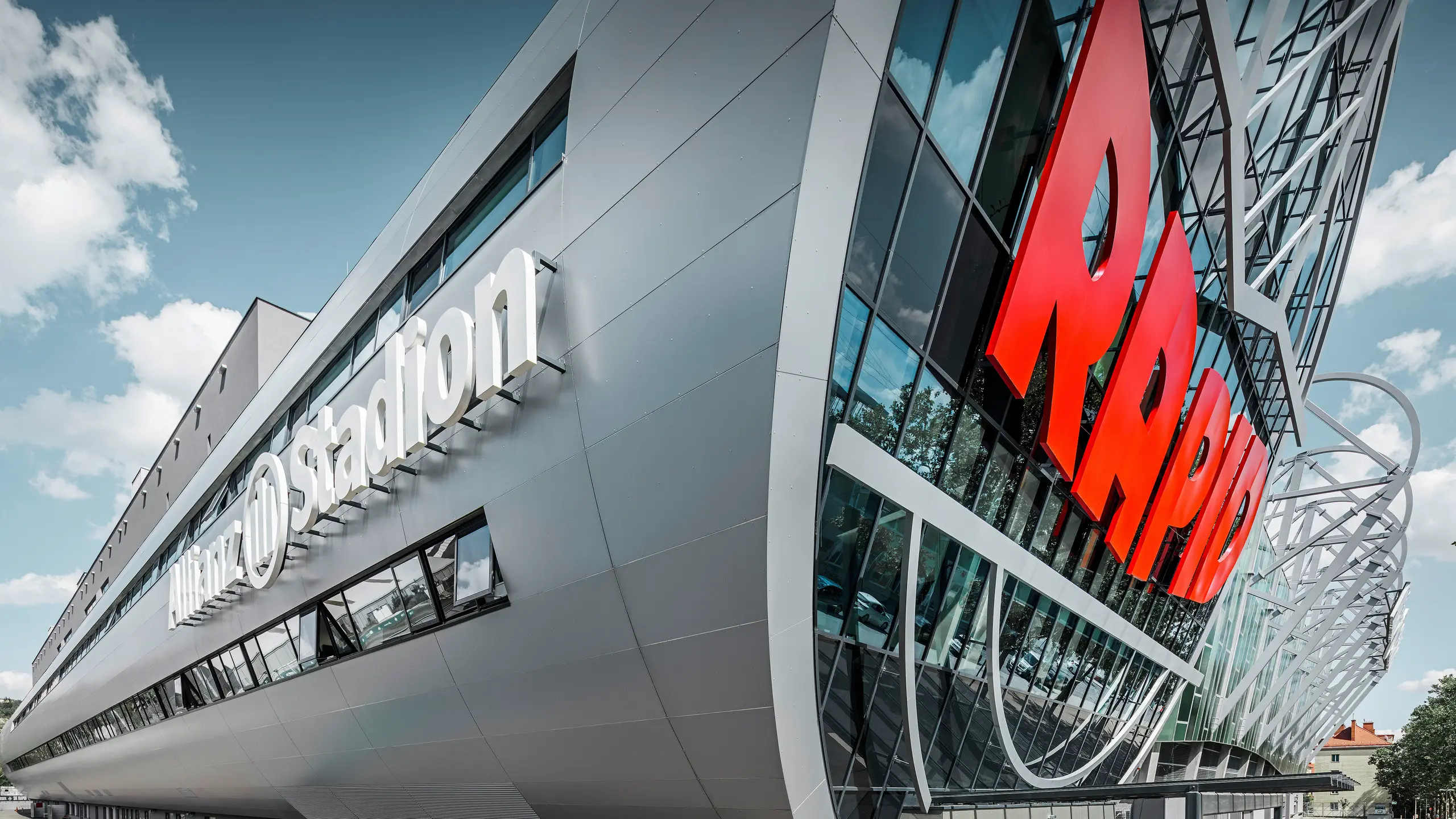 Krupni plan fasade stadiona Alijanc, doma FK Rapid Beč. Impresivni tunel u ulaznom delu prikazuje PREFA aluminijumske kompozitne panele u srebrnoj metalik boji. Ugao gledanja naglašava modernu arhitekturu. Jarko crveni natpis RAPID i čiste linije srebrnih panela daju tunelu vizuelno upečatljivu dinamiku i estetsku privlačnost. Na ovaj način, strast se rasplamsava kod svih fudbalskih navijača čak i pre nego što stignu na stadion.