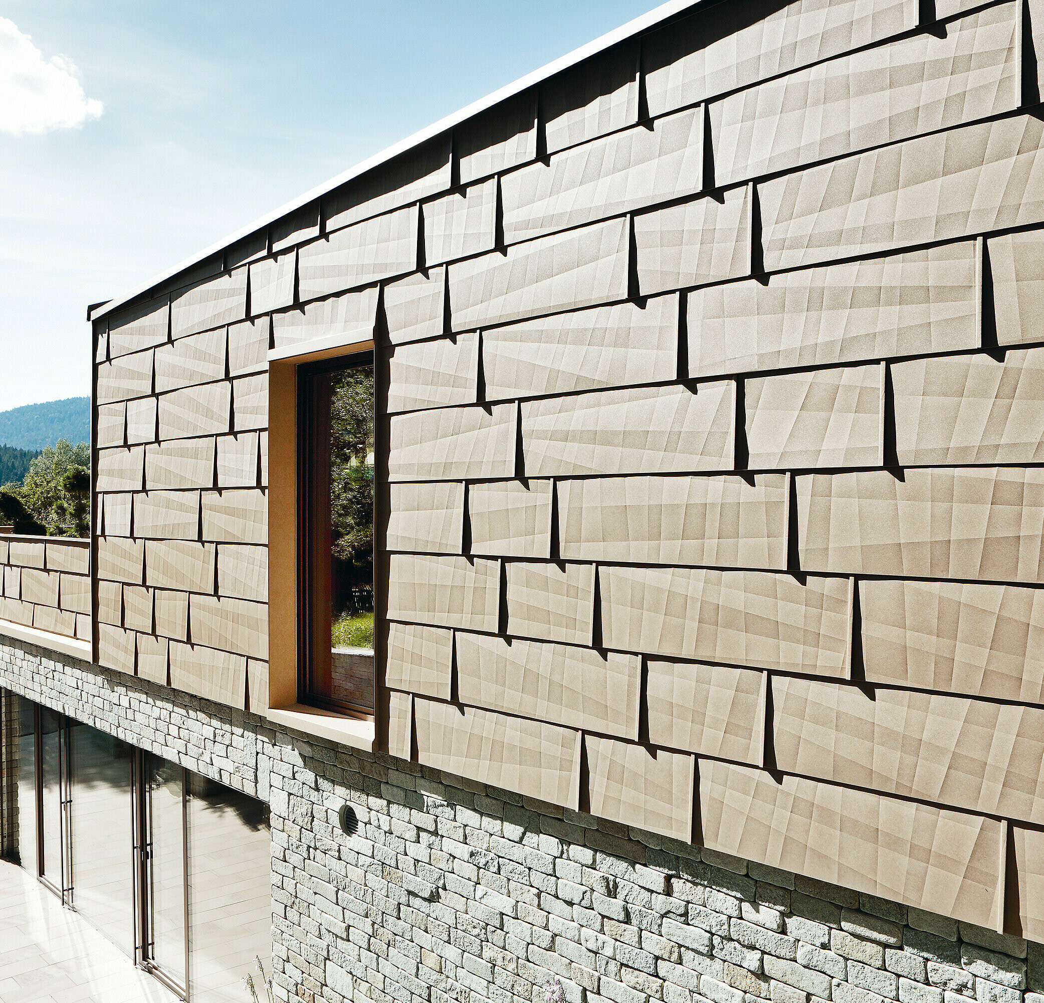 An dieser Außenfassade wurde das PREFA Fassadenpaneel FX.12 in P.10 Sandbraun mit einer Steinfassade im Untergeschoß kombiniert.