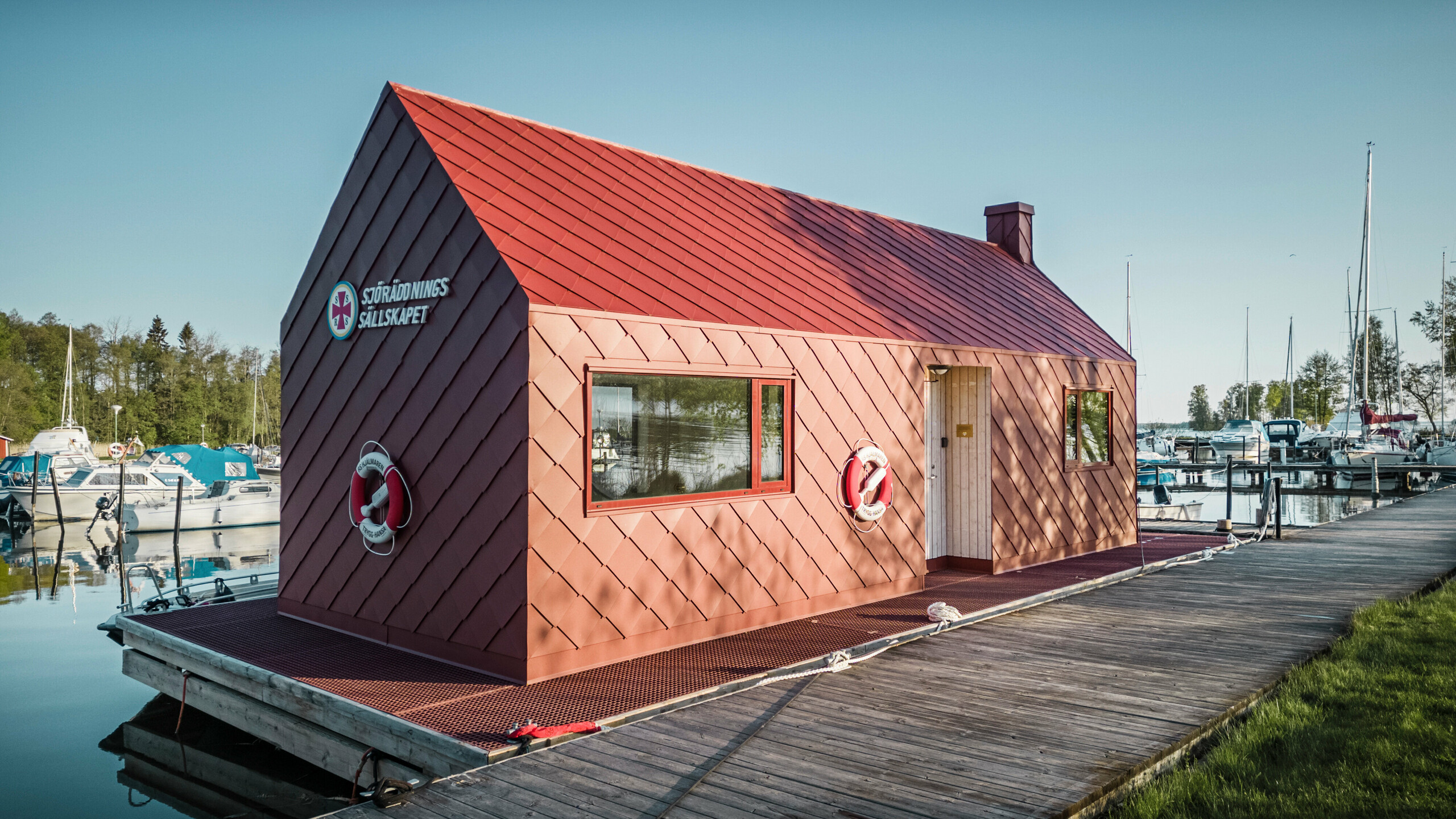 RS Hjälmaren rescue station on Sweden's fourth-largest lake Die Seenotrettungsstation am Hjälmaren See in Schweden, festgemacht an einem Holzsteg. Das markante Gebäude mit einer Fassade aus oxydroten PREFA Dach- und Wandrauten 29×29 steht stolz im Kontrast zu den weißen und blauen Booten im Hintergrund. Mit Rettungsringen ausgestattet und bereit für den Einsatz, vermittelt das Haus sowohl Sicherheit als auch einen starken maritimen Charakter in dieser friedvollen Seenlandschaft.