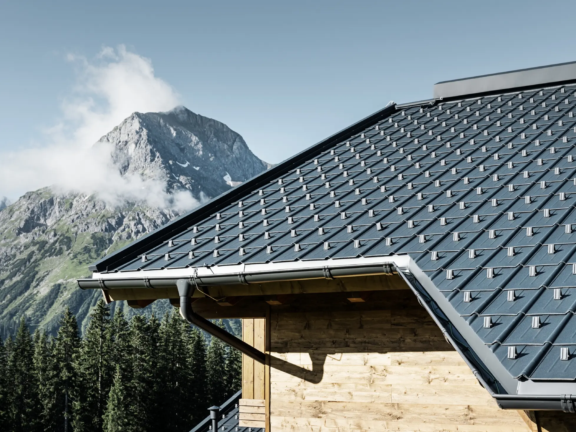 PREFA krovna ploča, primena u alpskom okruženju - krovna ploča u boji antracita postavljena na kolibu u naselju Lech am Arlberg, sa planinama u pozadini; PREFA oluk u boji antracita; drvena fasada
