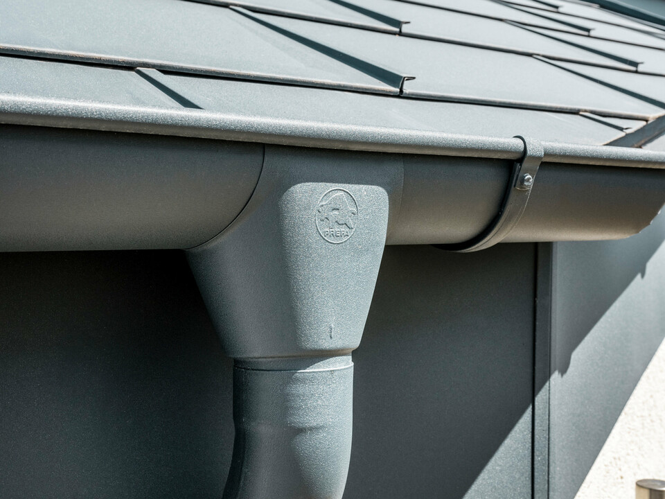 Nahaufnahme eines Hausdaches mit PREFA Dachplatte R.16 in Anthrazit sowie detailreicher Ansicht der farblich abgestimmten PREFA Dachrinne und Fallrohr aus Aluminium bei sonnigem Wetter.