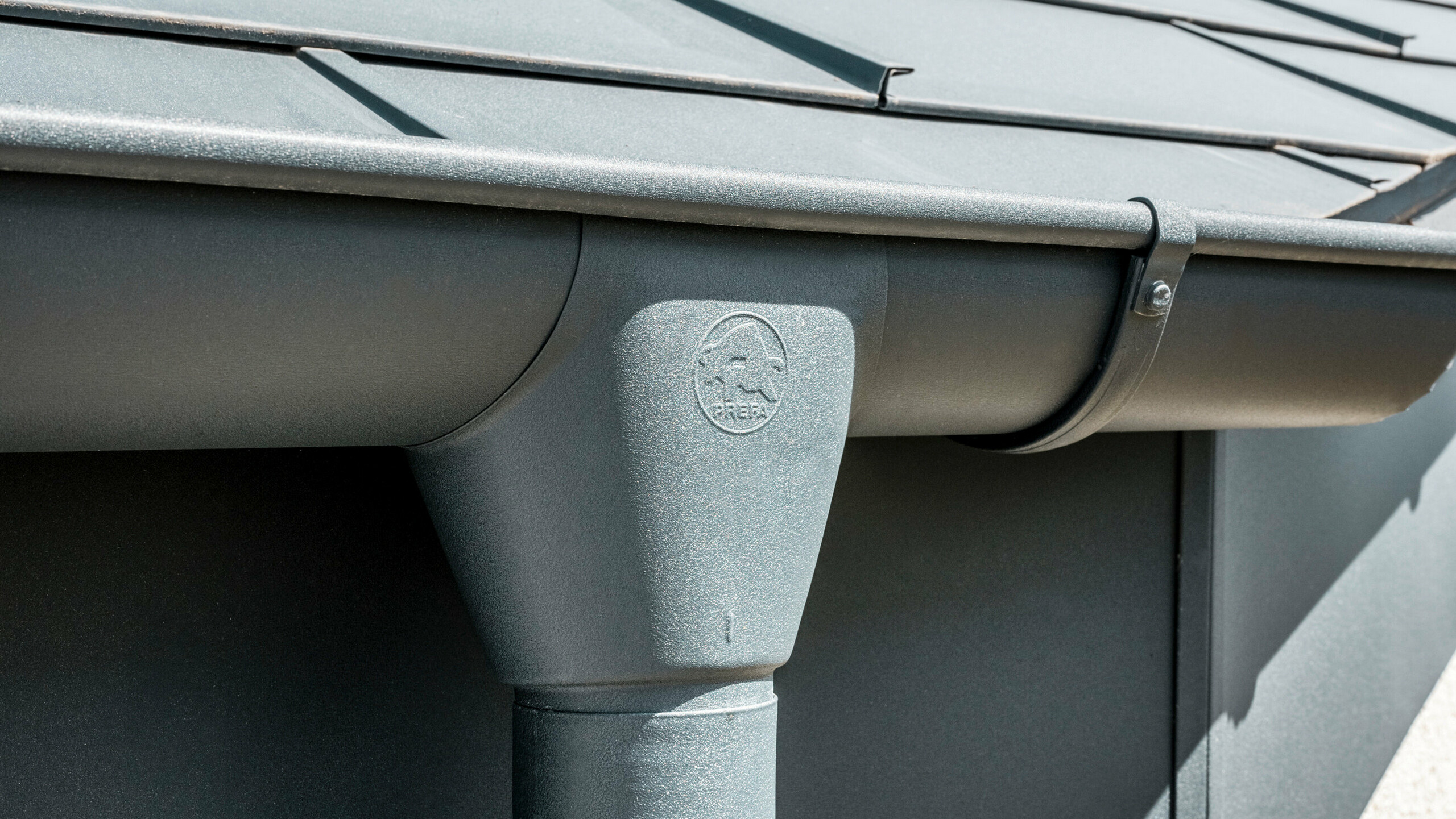 Nahaufnahme eines Hausdaches mit PREFA Dachplatte R.16 in Anthrazit sowie detailreicher Ansicht der farblich abgestimmten PREFA Dachrinne und Fallrohr aus Aluminium bei sonnigem Wetter.