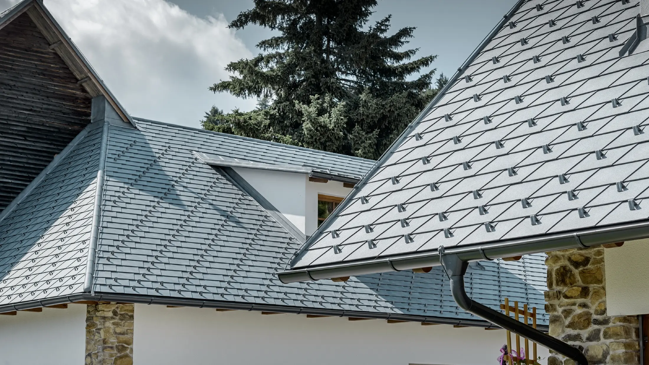 Detaljni prikaz aluminijumskog krovnog pokrivača proizvođača PREFA; krovne šindre u kamen sivoj boji sa aluminijumskim krovnim olukom u antracit boji proizvođača PREFA; u pozadini se vidi prozor sa izduženim krovom sa stojećim falcom. Fasada je bela sa ugrađenim kamenim elementima.